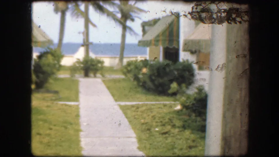 1945 Florida seaside resort view with a path, lawn, palms, white buildings, striped awnings, and blue water in the distance.