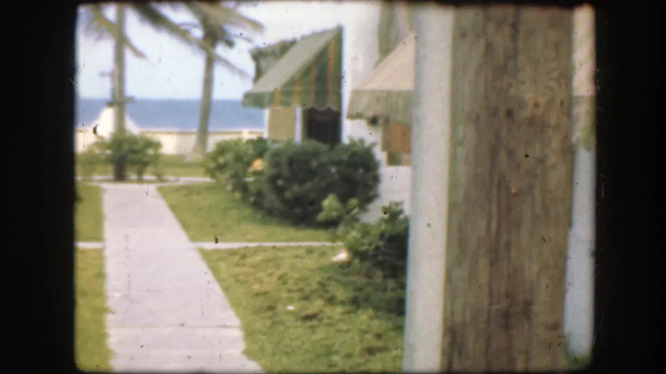 1945 Florida resort grounds from a visitor viewpoint, with a walkway, palms, awnings, ocean, and wooden post in the foreground.