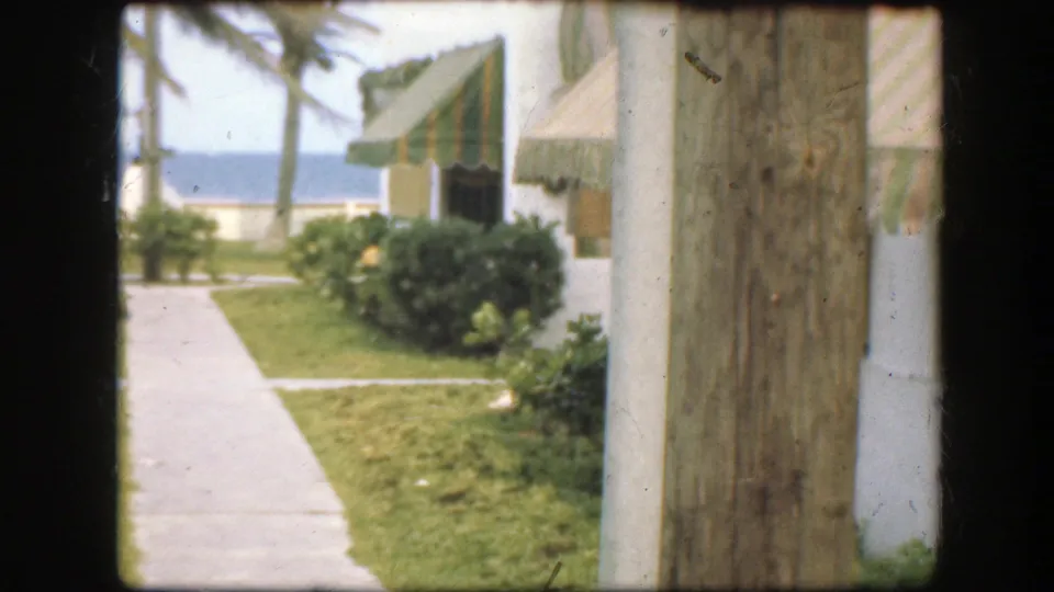 1945 Florida seaside resort exterior with a concrete path, trimmed grass, palms, striped awning, ocean, and wooden post at right.