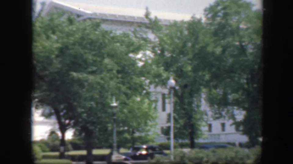 Washington DC home-movie view with landscaped trees, lamps and a dark parked car in front of a white building exterior.