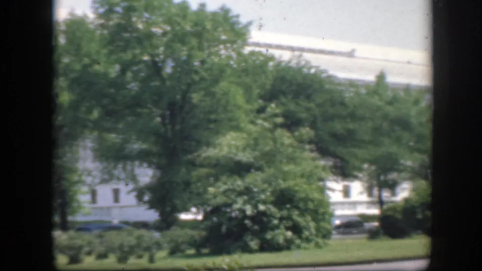 A soft 8mm Washington DC exterior shows a pale building partly screened by trees, with lawn and a dark parked car visible.
