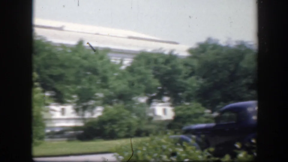 A handheld 8mm frame shows the Washington DC building area blurred by movement, with shrubs, lawn, and a dark car at the curb.