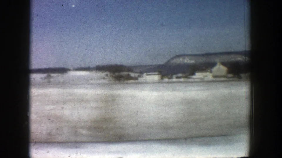 1961 Aspen road-view frame showing a wide snow-covered roadside, distant buildings, and a mountain ridge under clear sky.