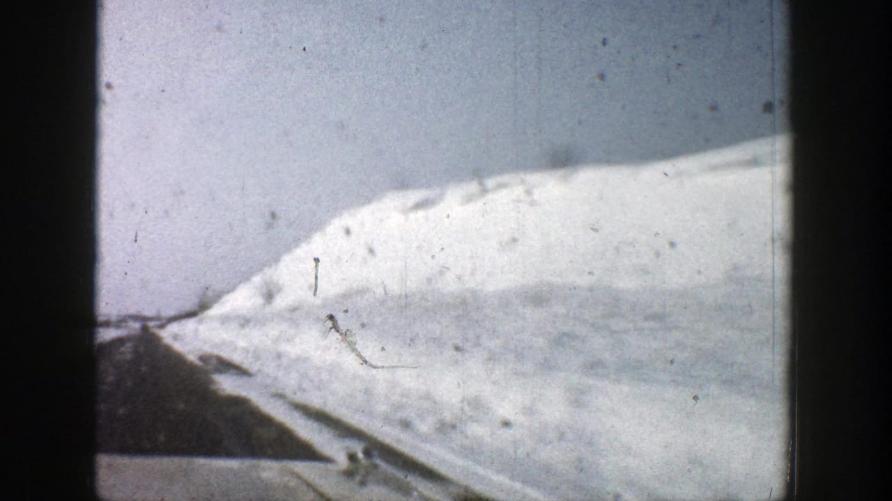 1961 8mm Aspen, Colorado frame showing a snowy hillside, road edge, and high roadside snowbank from a moving vehicle.