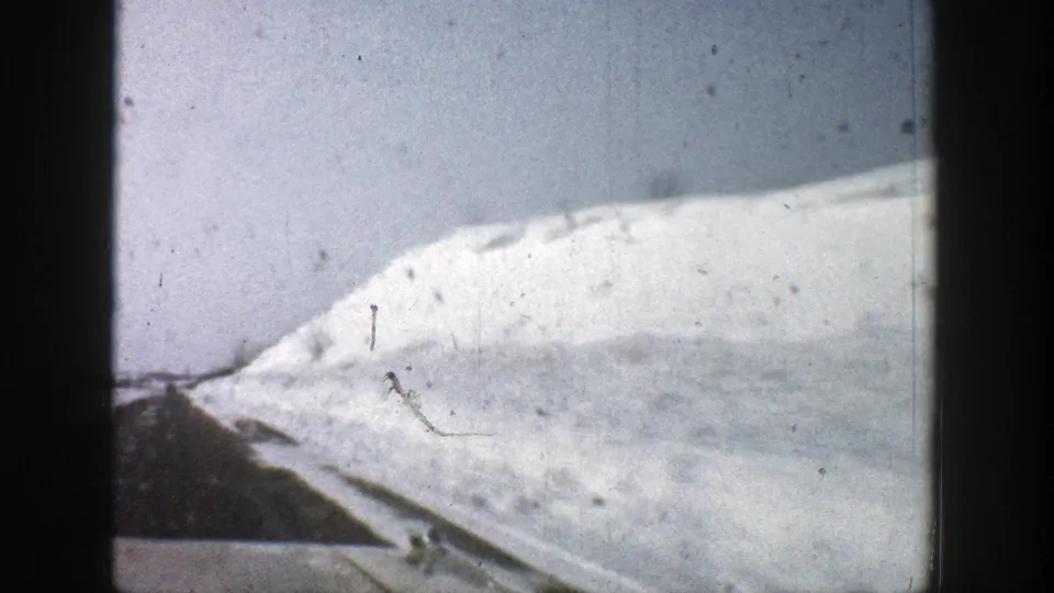 1961 8mm Aspen, Colorado frame showing a snowy hillside, road edge, and high roadside snowbank from a moving vehicle.