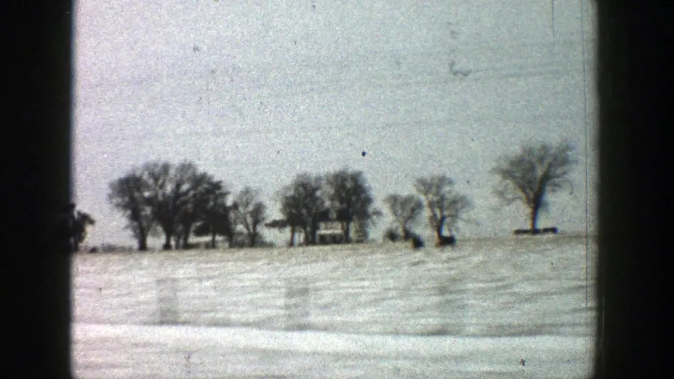 1961 8mm Aspen, Colorado frame showing a snow-covered field, bare winter trees, and a small distant farmhouse-like building.