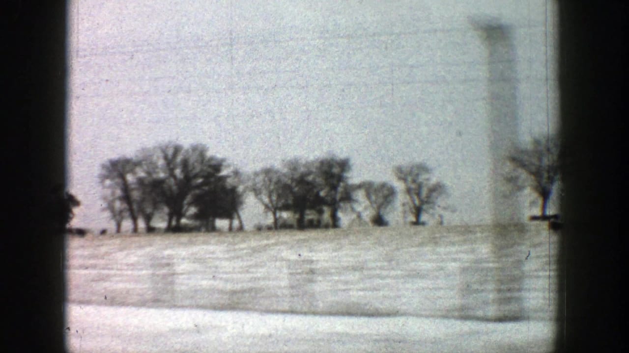 1961 Aspen, Colorado area home-movie view of a snowy rural field, bare trees, and distant buildings through a moving vehicle window.