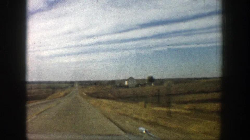 1961 rural road view near Aspen, Colorado, with fields, fence lines, distant structures, and a broad sky seen from a moving vehicle.