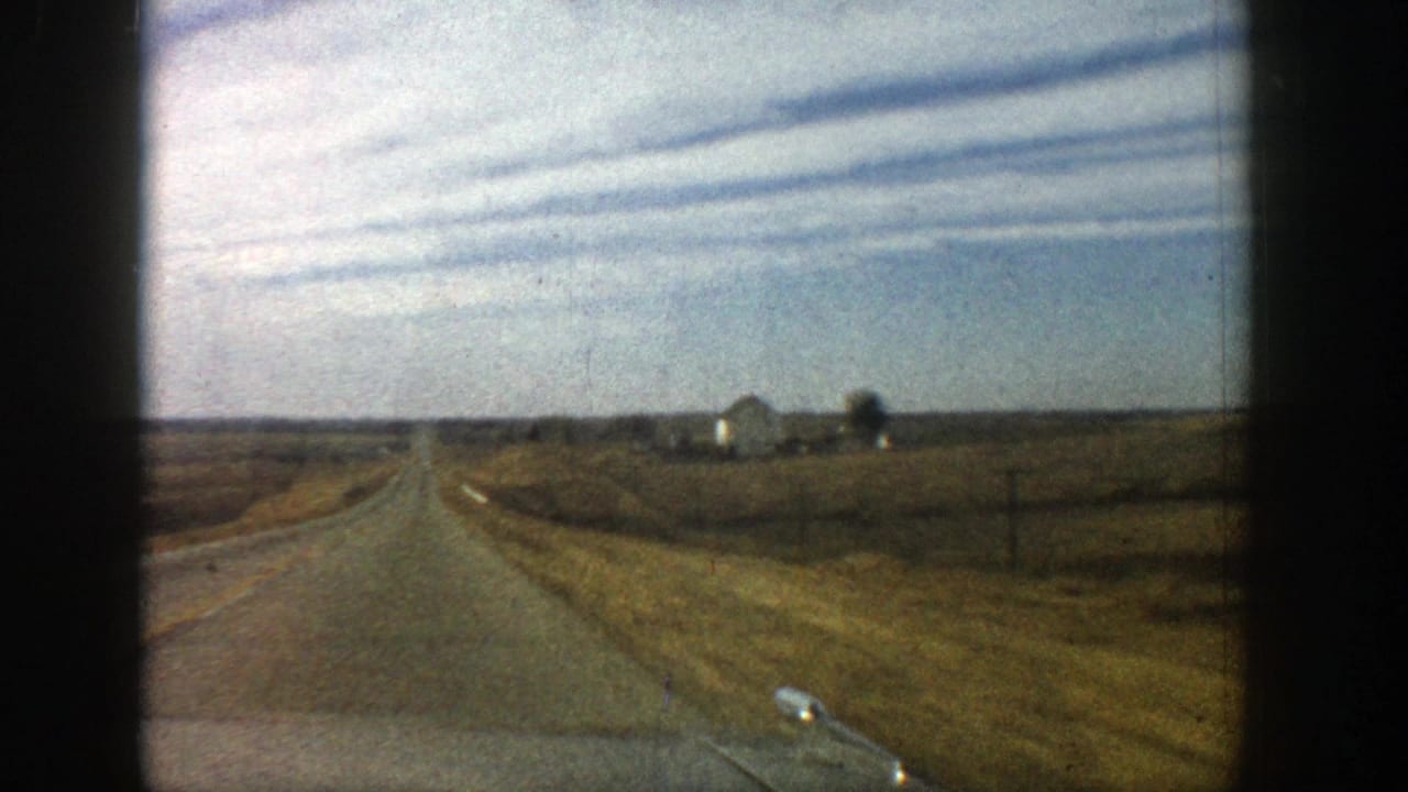 Paved rural road seen from a moving vehicle, with fields, utility poles, and distant farm buildings.