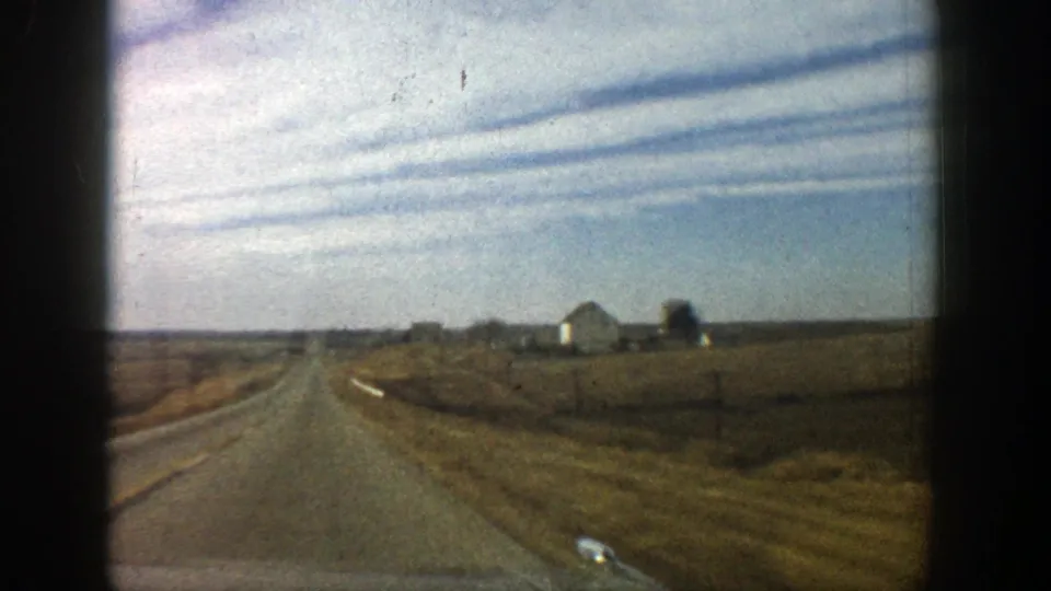 Vehicle-window view of a 1961 rural Colorado roadway, with farm structures set back from the road and broad fields under a pale sky.
