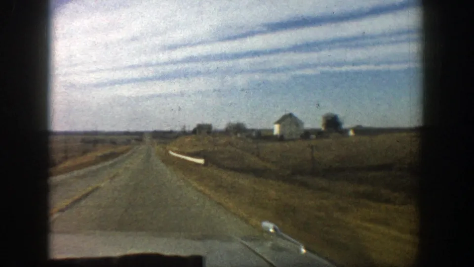 1961 8mm frame from a moving vehicle on a rural road near Aspen, showing fields, a white farm building, and low horizon line.