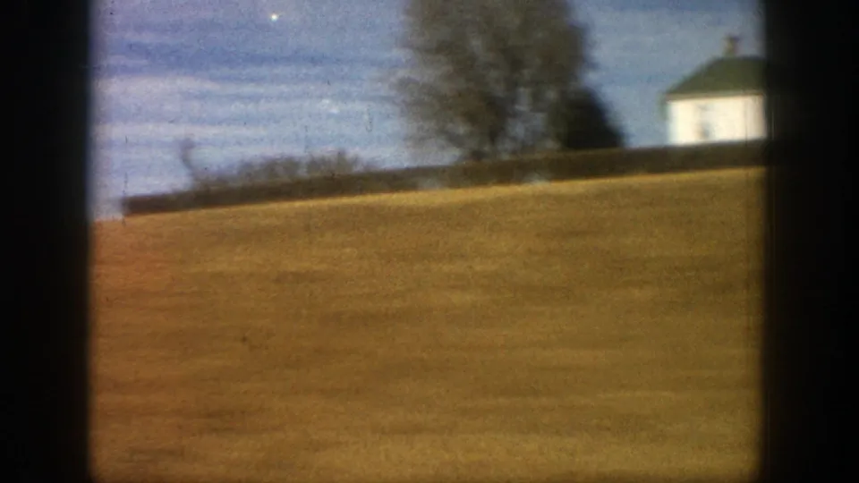 The moving 8mm camera passes a sloped rural field with a small white building and bare trees in Aspen, Colorado.
