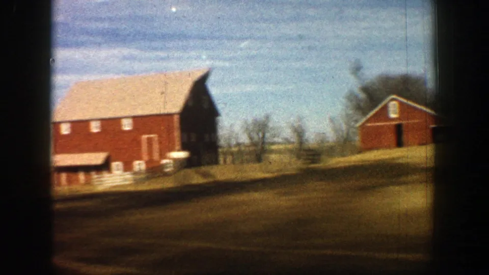 Red barn structures stand along an open rural field in this 1961 Aspen, Colorado 8mm driving shot.