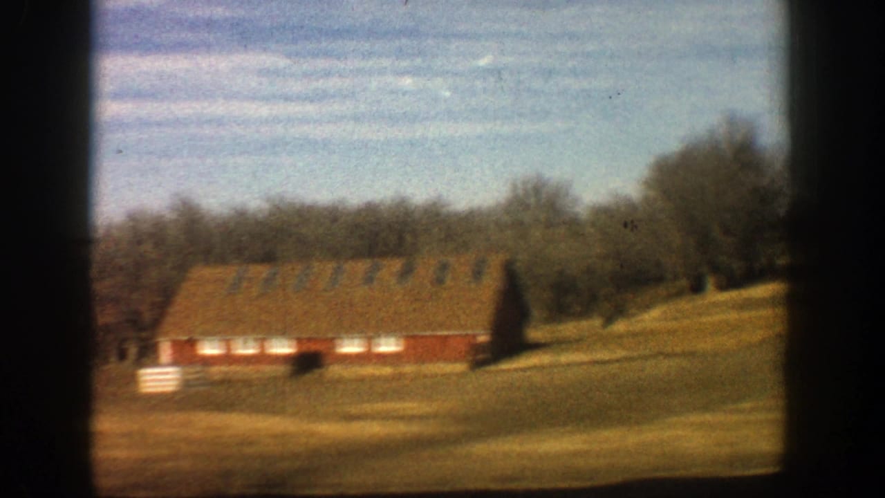 Long red farm building with white trim on a grassy slope under a blue sky.
