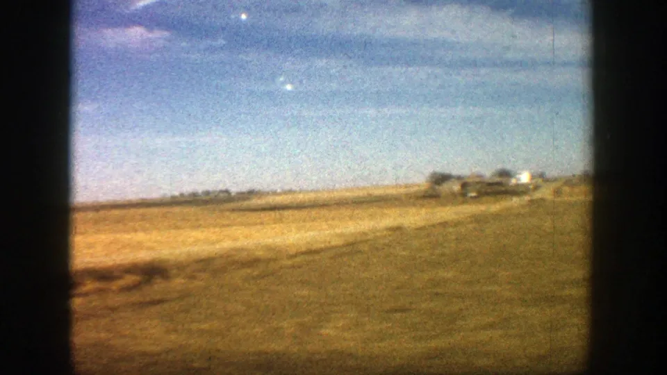 1961 8mm driving view near Aspen showing a road edge, yellow-brown fields, low structures, and soft clouds overhead.