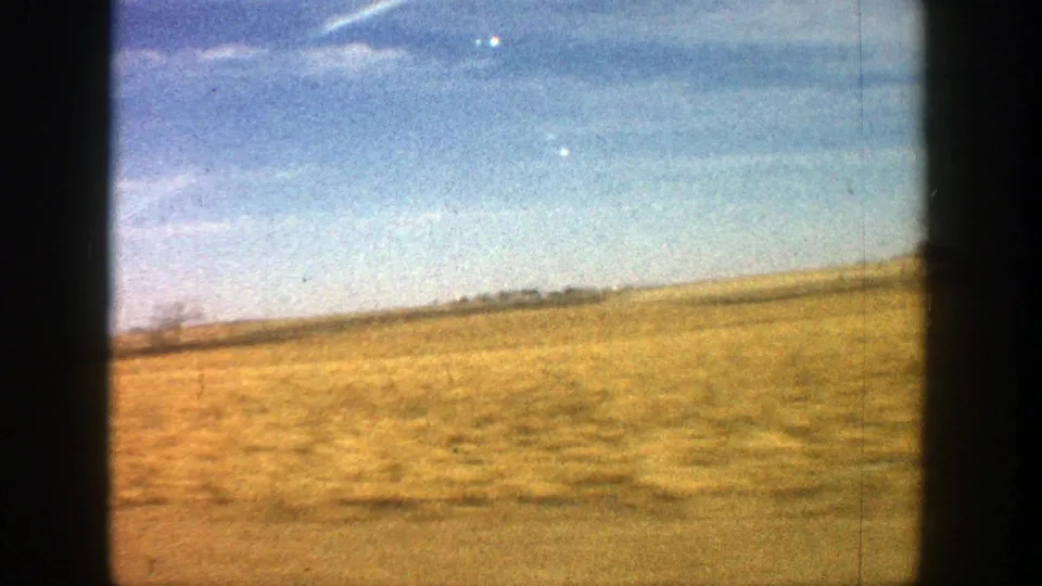Late-frame 1961 rural Aspen driving shot with motion-softened fields, a distant horizon, blue sky, and visible 8mm film texture.