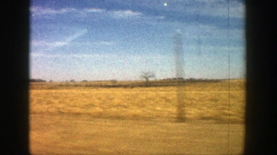 1961 Aspen-area roadside view with dry open ground, faint distant trees, and a bright sky.