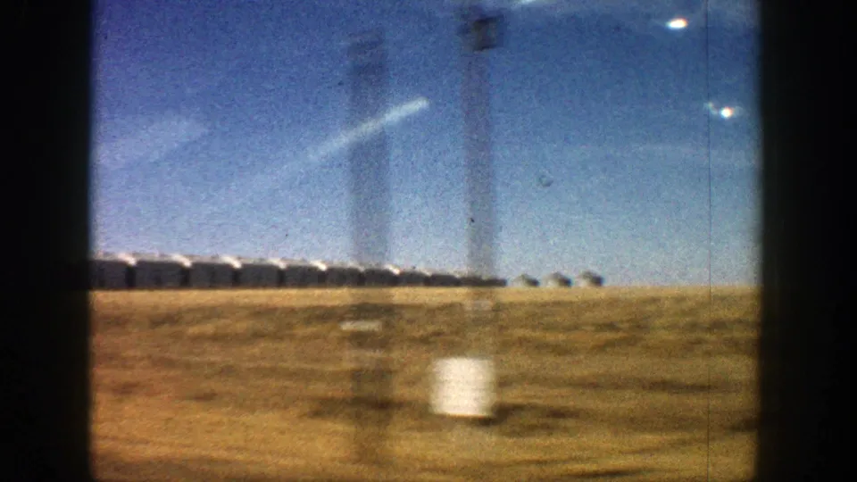 1961 Aspen-area 8mm frame with white roadside buildings beyond a dry field, seen from a moving car.