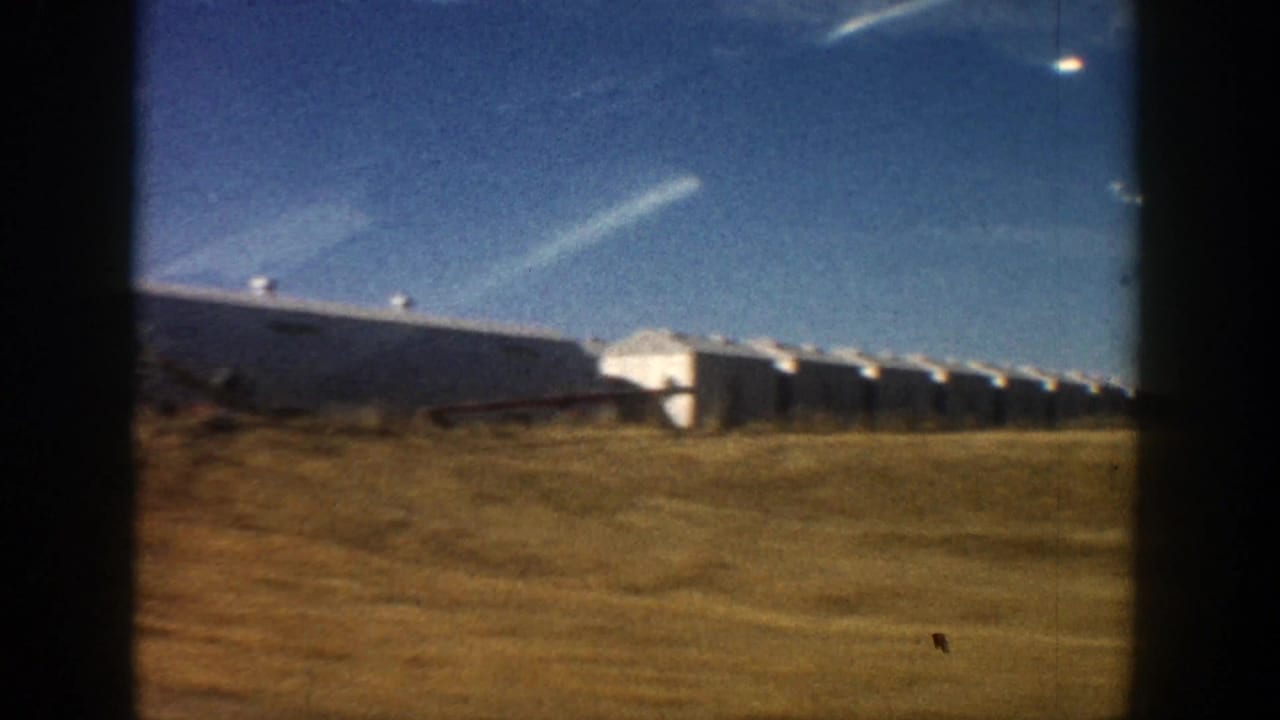 1961 Aspen-area 8mm frame showing long white farm buildings beyond a grassy roadside under a blue sky.
