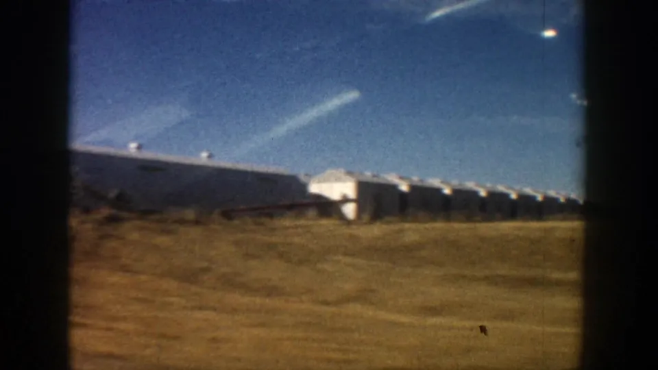 1961 Aspen-area 8mm frame showing long white farm buildings beyond a grassy roadside under a blue sky.