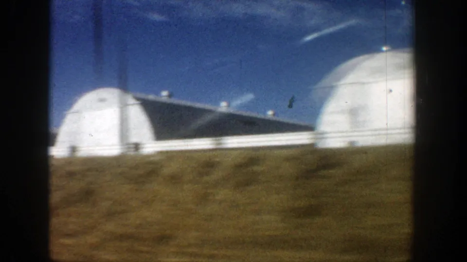 1961 Aspen 8mm view of rounded white agricultural structures and a long building seen while passing by car.