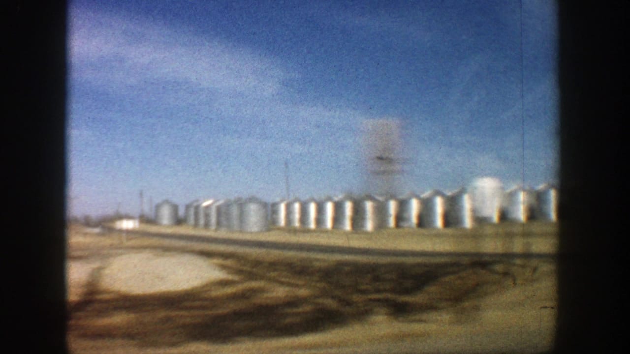 1961 Aspen, Colorado footage appears to show a long row of silver storage tanks in a tan field, framed by dark vehicle or window edges.