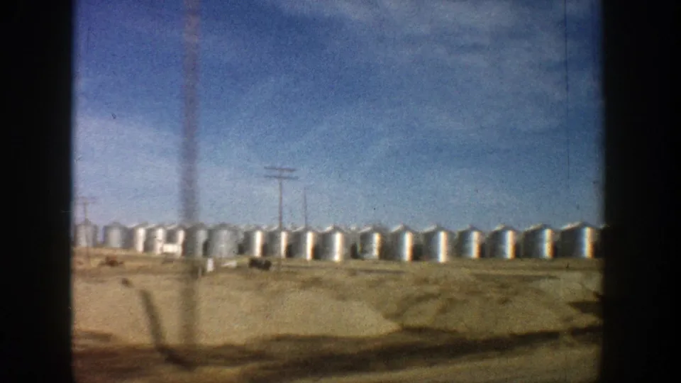 A moving 8mm view in Aspen, Colorado shows silver storage tanks, utility poles, and open tan ground under a bright sky.