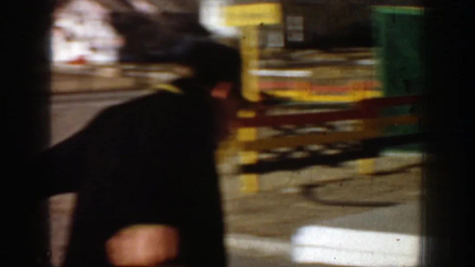 A blurred 1961 home-movie frame shows a man in dark clothing near a roadside facility or counter, with signage and structural framing visible.