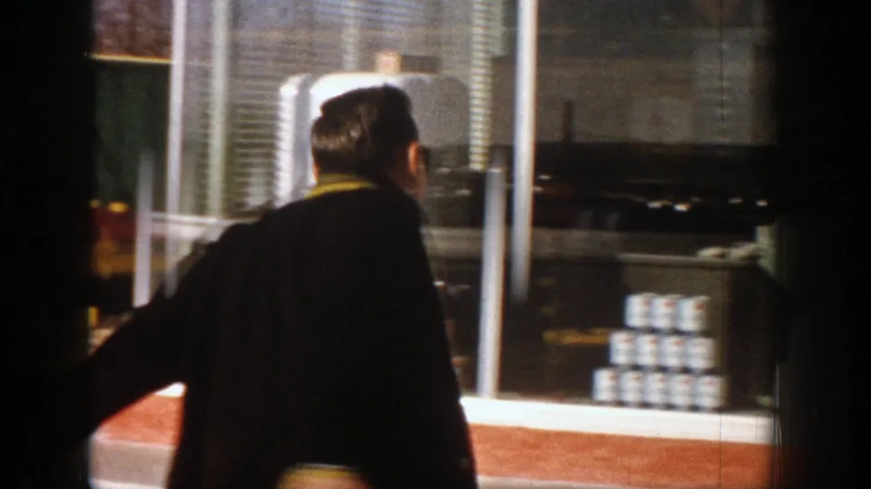 A man with a yellow collar is seen from behind at a counter or service window, with shelves, stacked containers, and exterior tanks visible.
