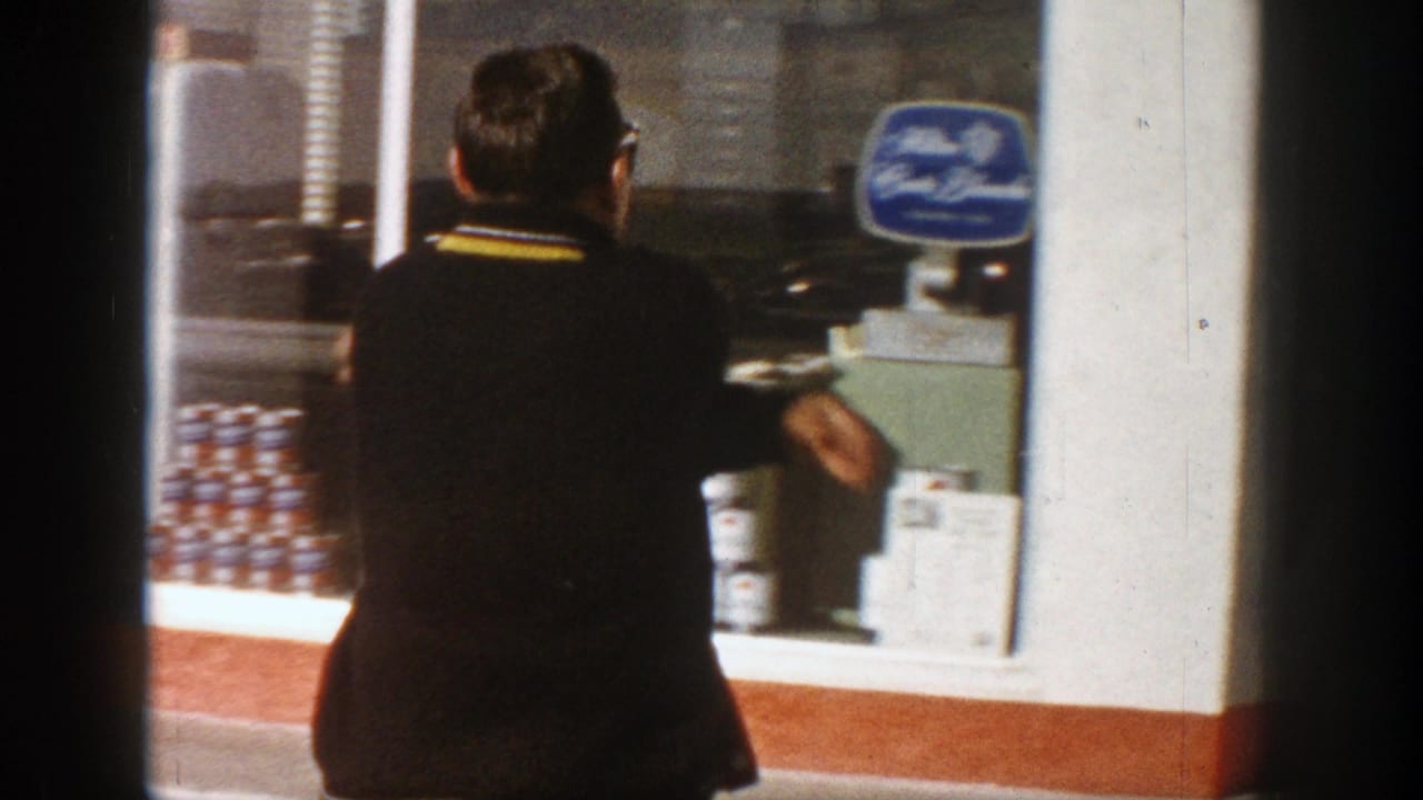 Adult man in a dark sweater stands at an Aspen storefront window in 1961, near stacked beer cans and a blue sign.