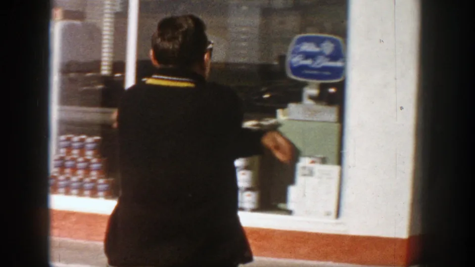 Adult man in a dark sweater stands at an Aspen storefront window in 1961, near stacked beer cans and a blue sign.