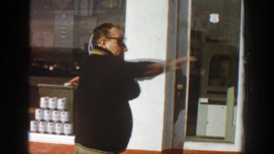 Adult man in sunglasses points along an Aspen storefront window in 1961, with stacked Miller High Life cans visible behind glass.