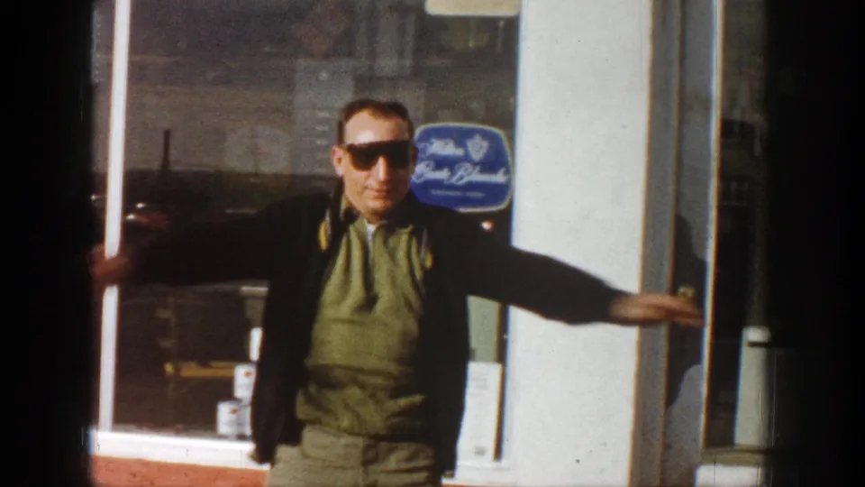 Adult man in sunglasses stands in front of an Aspen storefront in 1961 with his arms spread near the glass door and window display.