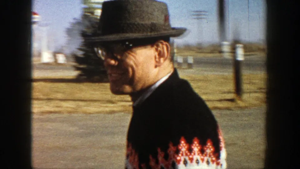 Adult man in a fedora and patterned sweater smiles outdoors in Aspen, Colorado in 1961, with a street and open background behind him.