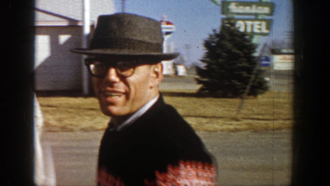 In 1961 Aspen, Colorado, a man in a fedora, glasses, and patterned sweater smiles while walking near a street and sign.