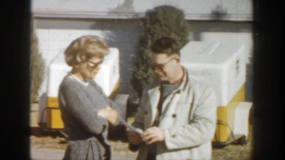 A woman in a gray dress and a man in a light jacket converse near white-and-yellow trailers in 1961 Aspen, Colorado.