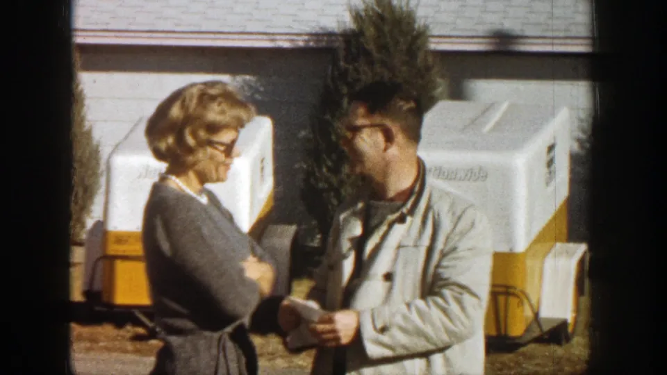 The woman and man continue their sidewalk conversation near trailers and a small tree in a 1961 Aspen street scene.