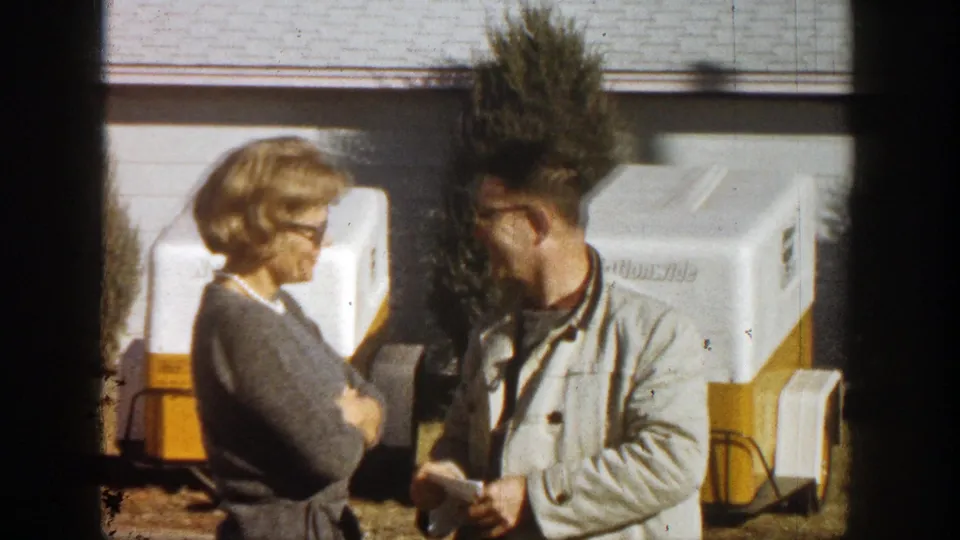 In supported 1961 Aspen home-movie footage, a woman and man stand near white and yellow coolers outside a house.