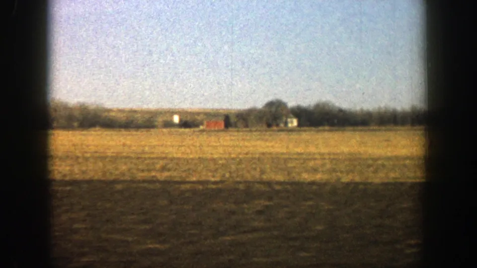 1961 8mm frame showing flat rural farmland with small distant farm buildings, including a red structure, against a low tree line.