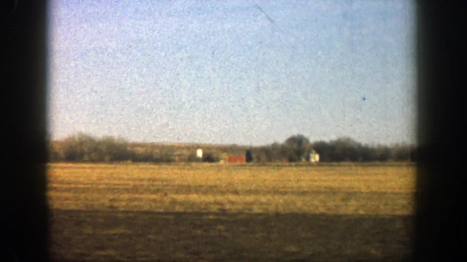 1961 rural vehicle-window view showing a broad field, distant farm buildings, a low tree line, and clear blue sky.