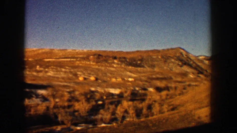 1961 Aspen, Colorado frame showing a rocky, dry hillside, sparse shrubs, blue sky, and warm 8mm color from a moving road view.