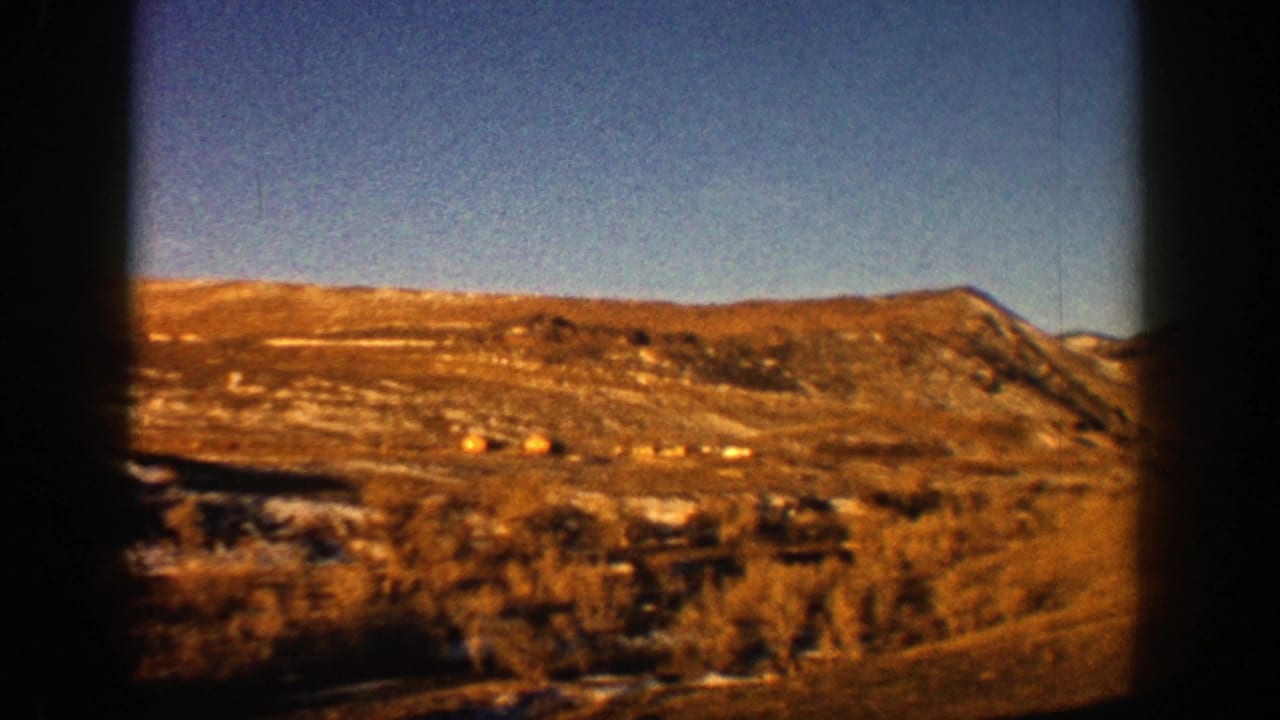 1961 Aspen-area 8mm road-view frame showing dry hillsides, distant mountains, and a clear blue sky from a moving vehicle.