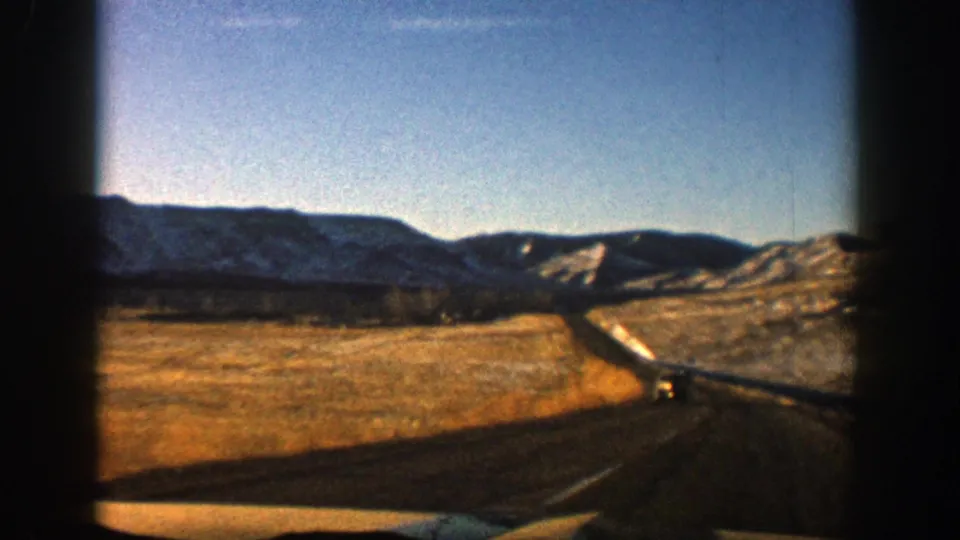 1961 Aspen-area highway view with a paved road curving through open terrain, patchy snow, and low mountain ridges.