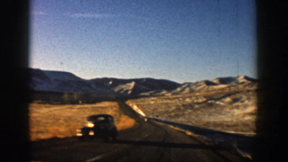 A black mid-century car passes on the highway in 1961 Aspen-area home-movie footage, with snowy hills and mountains beyond.