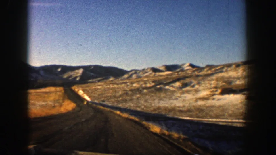 1961 Aspen-area 8mm frame showing a highway receding through snowy, rugged hills under a bright blue sky.