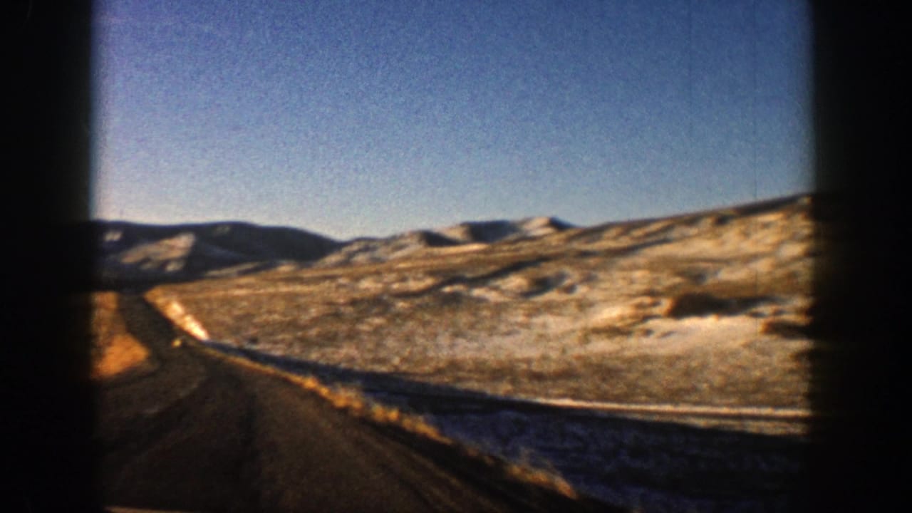 1961 Aspen, Colorado 8mm frame showing a road edge, open valley, distant hills, snow patches, and blue sky from a moving car.