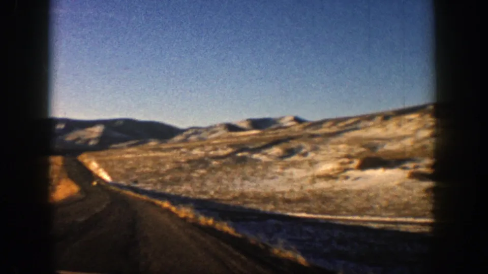 1961 Aspen, Colorado 8mm frame showing a road edge, open valley, distant hills, snow patches, and blue sky from a moving car.