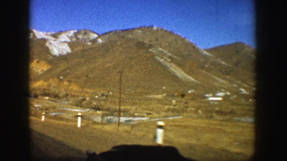 1961 Aspen, Colorado road scene with white roadside posts, utility pole, scattered distant structures, and snow-dusted mountain slopes.