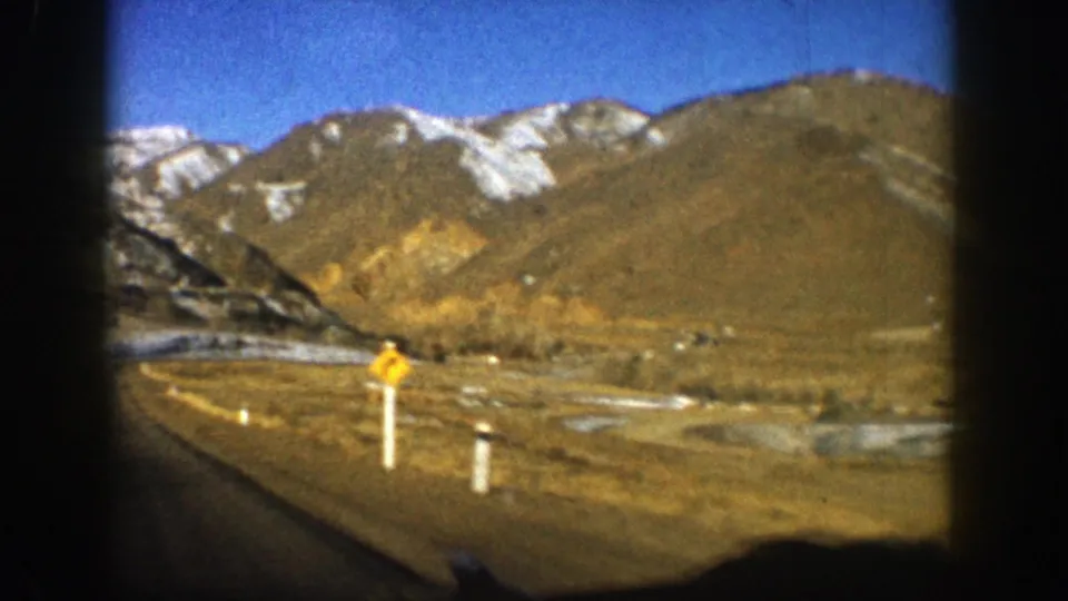 1961 Aspen, Colorado frame with a yellow curve sign, roadside posts, winding road, and snow-marked mountains in clear daylight.