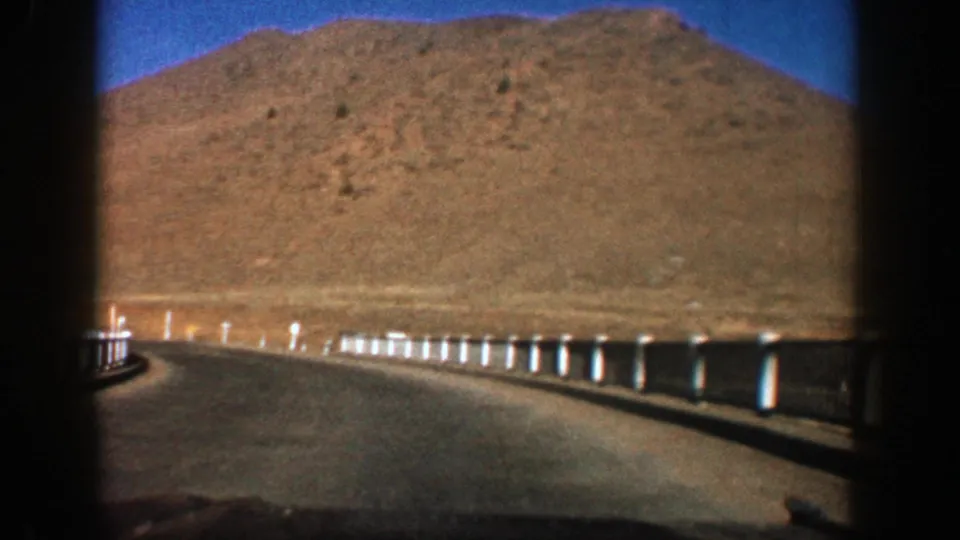 Aspen, Colorado 8mm travel frame from 1961 with guardrail posts, curved pavement, and a sunlit arid hillside.